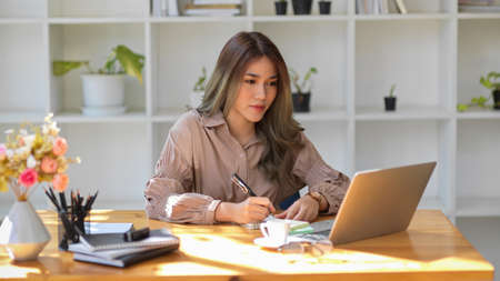 Attractive Asian female university student taking an online class, lecturing on her notepad in a library workspace.の写真素材