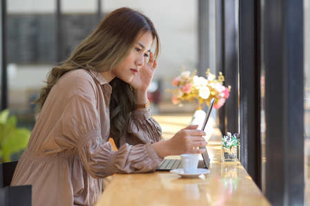 Stressed woman working on portable tablet touchpad in a coffee shop co-working space. A female freelancer is concerned about her career.の写真素材