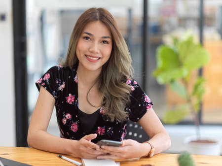 Beautiful Asian millennial woman sitting in the coffee shop, holding smartphone and smiling to camera.の写真素材