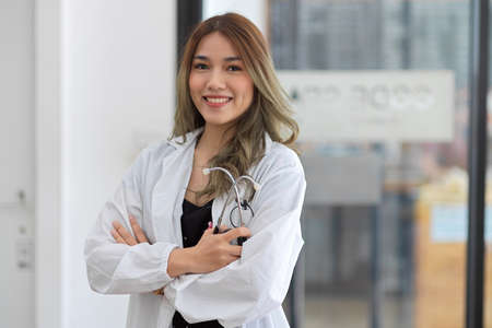 A professional and attractive Asian young female doctor in a white coat stands with her arms crossed and hold stethoscope in the hospital hallway.の写真素材