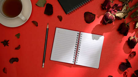 Stylish red workspace background with rose petals, tea cup, pencil and empty notebook page mockup. top viewの写真素材