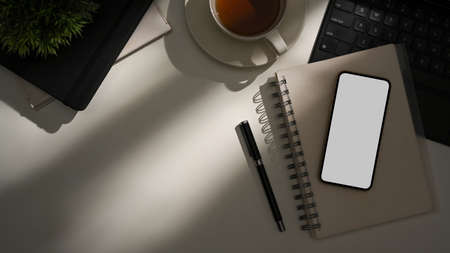 A modern hipster workspace with a daylight shadow on the table, office supplies, a coffee cup, a smartphone blank screen mockup, and a copy space.の写真素材