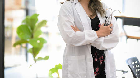 Cropped image of a female doctor in a white gown standing in her office with her arms crossed and holding a stethoscope.の写真素材