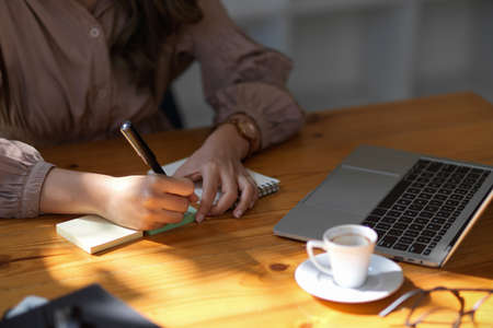 Cropped shot of female college student taking note on blank notebook while searching information on laptop in library or coffee shop.の写真素材