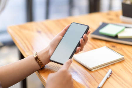 Close-up image, A female holding a smartphone white screen mockup over wooden table. Female searching something on internet.の写真素材