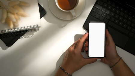Minimalist home office workspace desk. Woman hands holding smartphone mockup over desk with sunlight shadows. Flat lay, top view.の写真素材