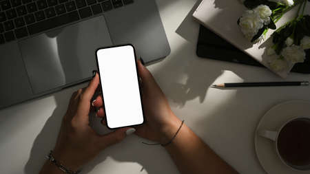 Top shot, Modern hipster workspace with female hand holding a smartphone blank screen mockup and sunlight shadow.の写真素材