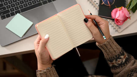 Woman hands in comfort sweater writing on notebook or diary, accessories and laptop on desk. Top view and copy spaceの写真素材