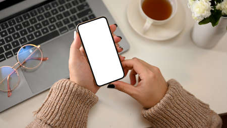 Minimal cozy female office desk with female in cozy brown sweater holding mobile phone white screen mockup. close-upの写真素材