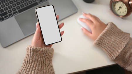 Above view, Woman in cozy sweater holding smartphone mockup with white screen over her office desk.の写真素材