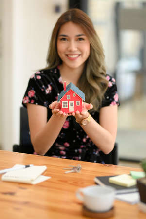 Portrait of a beautiful asian woman holding a stylish house model over office desk. Residential investment, Property purchase, House construction.の写真素材