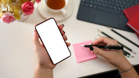 A female hand holding a smartphone blank screen and taking notes on sticky note on modern feminine office desk. top viewの写真素材