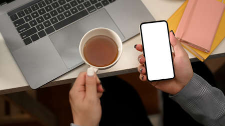 Top view, Businesswoman hands holding a teacup and smartphone white display mockup over modern office desk.の写真素材