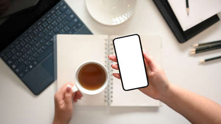 A modern minimal white workspace or female office desk with a female hands holding a teacup and smartphone blank screen mockup. top viewの写真素材