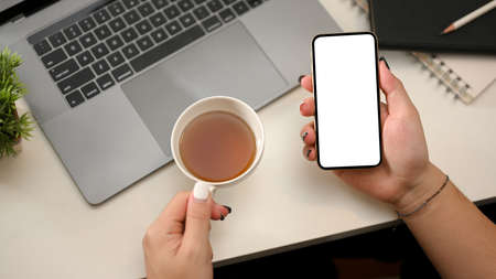 Top view, Modern office table with woman hands holding teacup and smartphone blank screen mockup.の写真素材