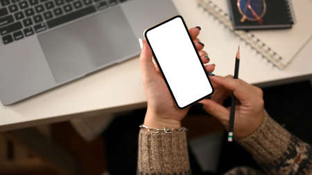 Cropped shot, top view of a female in cozy sweater hands holding a mockup of smartphone white screen...