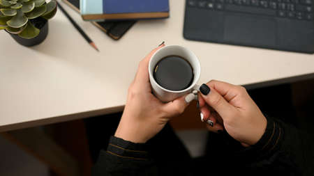 Cropped image, A female in black sweater holding a hot coffee cup over her modern office desk. aboveの写真素材