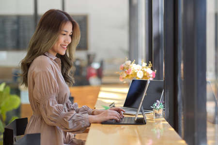 Side view, Beautiful Asian businesswoman working on tablet touchpad, typing on wireless keyboard at cafe coffee table.の写真素材