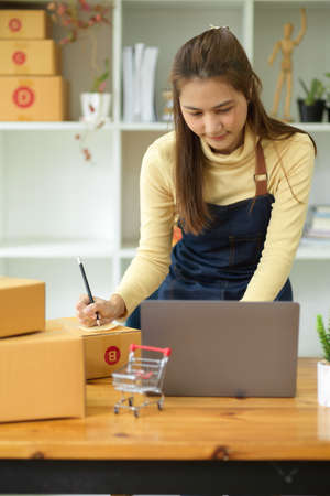 Portrait, Beautiful female small businesses owner, entrepreneur write the address on receipt box and check online orders on laptop computer.の写真素材