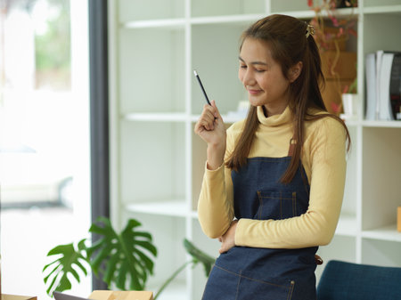 A pretty asian female in jean apron thinking a plan for her online business in her home office. SME entrepreneur and business concept.の写真素材