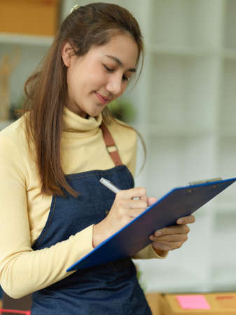Portrait of a beautiful asian female waitress taking a food and drink orders on clipboard paper. Friendly server wears apron and takes orders in restaurant.の写真素材