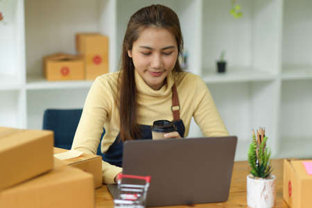 Beautiful Asian young female online shopping business owner working on laptop computer with parcel box in her home office. online selling, SME business.の写真素材
