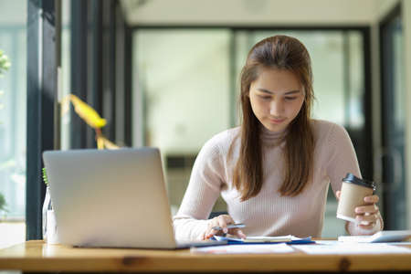 Professional Asian young businesswoman financial advisor sipping coffee while recheck the report and work on project alone in office.の写真素材