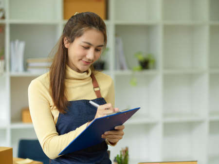 A beautiful asian young female business start-up writing the customer orders on clipboard. E-commerce business people.の写真素材
