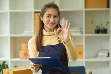 An attractive female online business owner, entrepreneur doing an OK hand sign and holding a clipboard in the office. E-commerce business, online shopping business.の写真素材