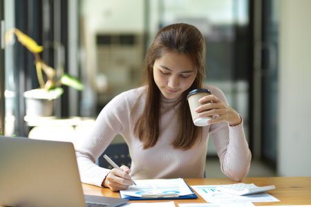 Charming Asian female accountant checking economic paper report, making remarks on the documents at workplace.の写真素材