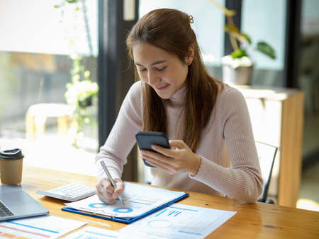Concentrated Asian young businesswoman holding smartphone, analysing financial report, working in modern office workplace.の写真素材