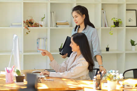 A successful and professional asian female manager briefing and checking her assistant's work on pc desktop screen in the office. Businesswomen conceptの写真素材