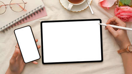 Stylish feminine workspace with a female hands holding smartphone and using stylus pen to draw on digital tablet touchpad. devices white screen mockup. top view, flat layの写真素材