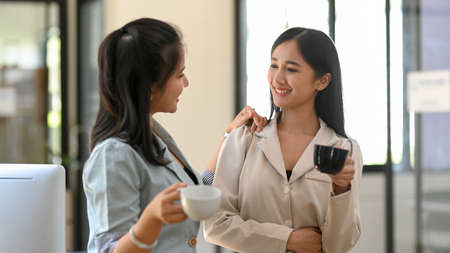 Two female colleagues are having a chill talk while sipping coffee on their coffee break in the office's snack room. Friendly businesswomen concept.の写真素材