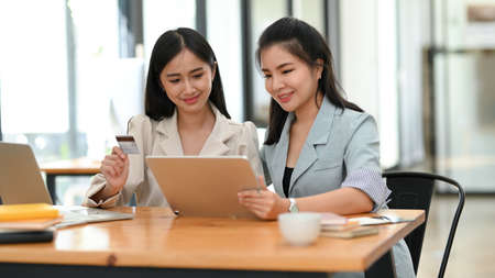 Two beautiful asian businesswomen sits in a modern cafe, uses portable digital tablet to pay their bills. Cashless payment conceptの写真素材