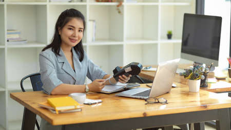 An Asian businesswoman holding a credit card and payment terminal machine at her office desk.の写真素材