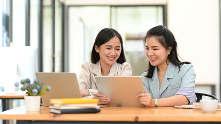 Two Asian young female business interns or trainees relax while watching some funny video on their tablet screens together.の写真素材