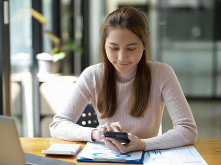 Positive asian young female accountant using a smartphone at her office desk. Woman relaxing on her mobile application.の写真素材