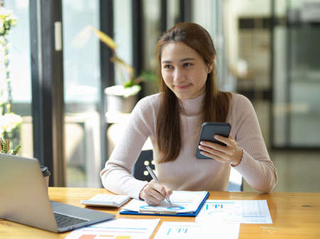 Pretty and charming female worker holding a smartphone while working on her paperwork and looking at laptop screen in the office.の写真素材