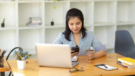 Female manager sits in modern office, paying her online shopping with credit card on smartphone.の写真素材
