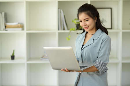 Professional asian female manager standing in office and holding an open laptop computer. Female...