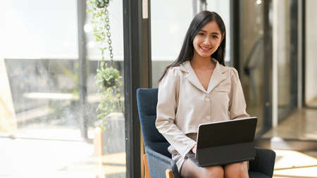 Attractive asian female sits on armchair and uses portable tablet computer in modern office space.の写真素材