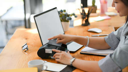 Attractive female banker swiping a credit card on payment terminal machine on her office desk. contactless technology, urban lifestyle.の写真素材