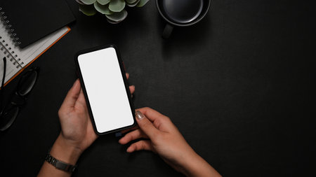 Overhead shot, A female hands holding smartphone white screen mockup over modern black workspace background.の写真素材