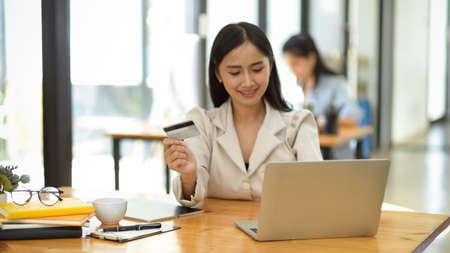 Attractive asian young female in white suit using portable laptop computer and holding a credit card. Online payment, online shopping conceptの写真素材