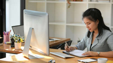Focused Asian businesswoman writing something on her personal notebook at her office workspace.の写真素材