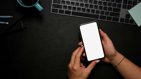 A black worktable with copy space and female hands holding smartphone white screen mockup over her office desk. top viewの写真素材