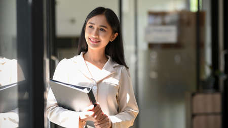 Charming asian young businesswoman or female employee holding binder folder and looking out the window with an inspired facial expression.の写真素材