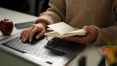 A female freelancer in cozy brown sweater looking something on her notebook and typing on laptop keyboard. Remote working conceptの写真素材