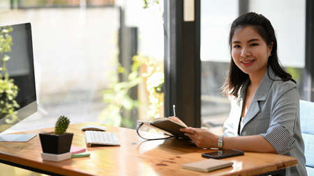 Professional female manager making a notes on her notebook, sitting at her desk and working on computer.の写真素材